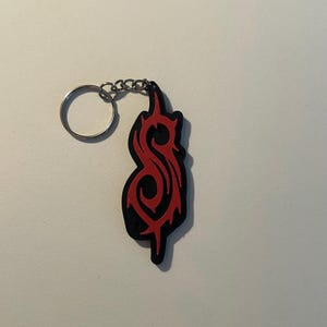 Könnte beinhalten: Ein schwarzer und roter Schlüsselanhänger mit einem stilisierten Design. Der Schlüsselanhänger hat einen silbernen Schlüsselring und eine Kette. Das Design ist ein rotes, flammenartiges Symbol auf schwarzem Hintergrund. Der Schlüsselanhänger ist wahrscheinlich aus Gummi oder einem ähnlichen Material.