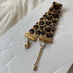 Può includere: Un braccialetto di perline con un motivo leopardato nero, marrone e oro. Il braccialetto ha chiusure in metallo color oro e una catena di estensione. Il testo "Shine.jwl" è visibile sul braccialetto.