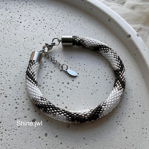 May include: A tubular beaded bracelet featuring a repeating black and white geometric pattern. The bracelet includes silver-toned metal end caps, a lobster claw clasp, an adjustable chain, and a small oval charm. The text Shine.jwl is visible.