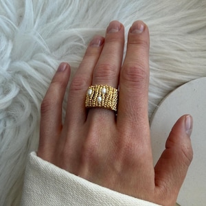 Puede incluir: Un anillo dorado con cuentas con tres perlas blancas. El anillo es ancho y cubre una gran parte del dedo. El anillo está en una mano con una manga de suéter blanca en el fondo.