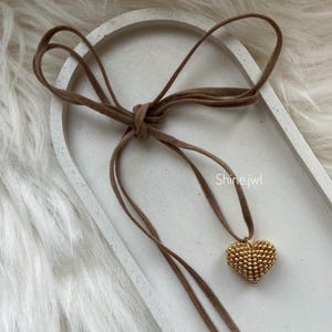 May include: A brown suede choker necklace featuring a gold beaded heart pendant. The necklace is tied in a bow. The text "Shine.jwl" is visible on the right side of the image.