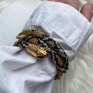 May include: A gold and black beaded snake bracelet with a gold snake head charm. The bracelet is wrapped around a person's wrist.