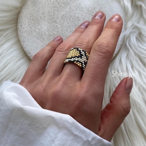 Peut inclure: Une bague perlée noire, dorée et blanche, portée sur une main. La bague présente un motif géométrique et est posée sur un fond blanc et duveteux. Le texte "Shine.jwl" est visible sur l'image.