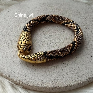 May include: A snake-shaped bracelet featuring a metallic gold head and tail with a flexible, tubular body crafted from small, alternating black and bronze beads. The text Shine.jwl is visible near the head of the snake-shaped jewelry piece.