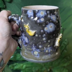 May include: A handmade ceramic mug with a dark, speckled glaze, adorned with raised yellow and white sculpted creatures. The mug features a sturdy handle and a light-colored interior, perfect for enjoying your favorite beverage.