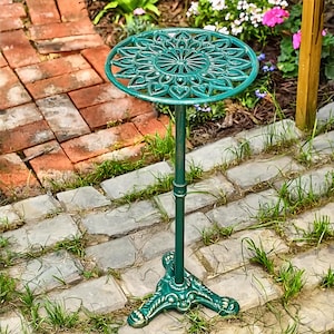 May include: A green, cast-iron garden table with a decorative floral design on the round tabletop. The table has a slender, vertical support and a three-footed base. The table is outdoors, set on a stone patio with grass and flowers.