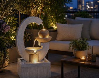 Puede incluir: Una fuente de agua para exteriores color piedra con un marco grande en forma de media luna, dos cuencos circulares apilados y un elemento superior esférico. La fuente incluye iluminación cálida integrada que ilumina el agua.