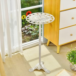 May include: A white metal plant stand with a decorative, openwork design on the circular tabletop. The stand has a slender, adjustable pole and a detailed, three-footed base. The stand is approximately 28 inches tall.