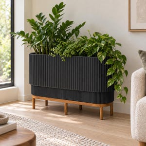 May include: A rectangular black planter with a vertical ribbed texture, supported by a light-toned wooden stand with four cylindrical legs. The planter contains a variety of green houseplants, including a tall Zamioculcas and a trailing pothos.