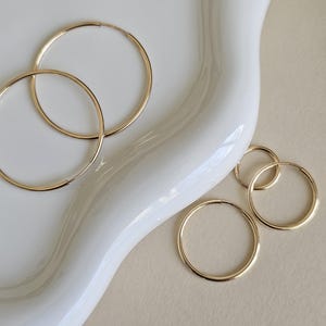 以下が含まれることがあります： 白い表面に置かれた、異なるサイズの3つの金のフープイヤリング