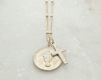 May include: A gold-toned necklace featuring a delicate satellite chain with two pendants: a round Miraculous Medal depicting a figure with rays of light and the text O MARY CONCEIVED WITHOUT SIN PRAY FOR US WHO HAVE RECOURSE TO THEE, and a small cross.