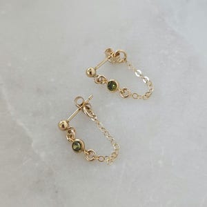 May include: A pair of gold-colored earrings with a chain and small green gemstones. Each earring has a post with a small ball, a green gemstone, and a chain that connects to a second green gemstone.