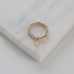 May include: A gold beaded ring with a small gold cross charm hanging from it. The ring is on a white marble surface.