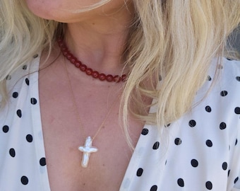 May include: A red beaded choker necklace and a gold chain necklace with a white cross pendant. The necklaces are worn over a white polka dot blouse.