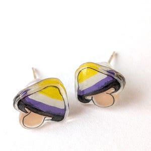 May include: Two mushroom stud earrings, each displaying the non-binary pride flag colours: yellow, white, purple, and black. The earrings have a clear, glossy finish and a post for pierced ears.