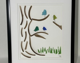 May include: Framed artwork featuring a tree with brown branches and four colorful birds perched on them. The birds are blue, green, and teal. Green grass is at the bottom. The artwork is in a black frame.