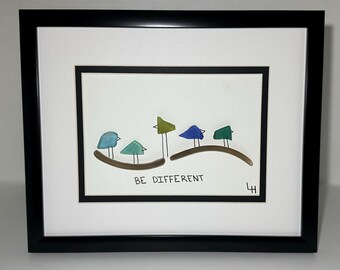 May include: Framed artwork featuring colorful bird-like shapes perched on a brown line. The birds are blue, green, and yellow. The phrase "BE DIFFERENT" is written below the birds in black text. The frame is black.