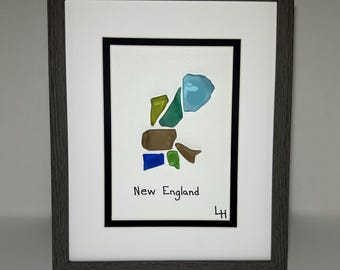 May include: Framed artwork featuring a map of New England created with colorful glass pieces. The artwork includes blue, green, brown, and yellow glass, with the words "New England" written below. The frame is gray.