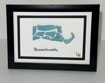 May include: Framed artwork featuring a map of Massachusetts crafted from light blue sea glass pieces. The state's name is written in black script below the sea glass design, set against a white background within a black frame.