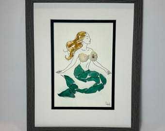 May include: Framed artwork of a mermaid. The mermaid has flowing golden hair, a shell top, and a green tail. The artwork is framed in a gray wooden frame with a white mat. The mermaid is drawn with black ink.