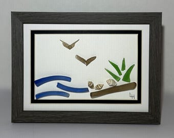 May include: Framed artwork depicting a beach scene. The art features blue glass waves, brown birds, green grass, and seashells on a white background. The frame is gray.