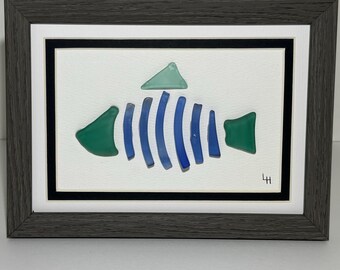 May include: Framed artwork featuring a fish design made from sea glass. The fish has blue and green glass pieces arranged on a white background. The frame is gray and the artwork is signed in the bottom right corner.