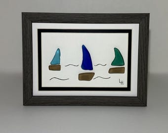 May include: Framed artwork featuring three sailboats with colorful sails. The sails are blue, green, and teal, with brown hulls, set against a white background. The artwork is framed in a dark gray wooden frame.