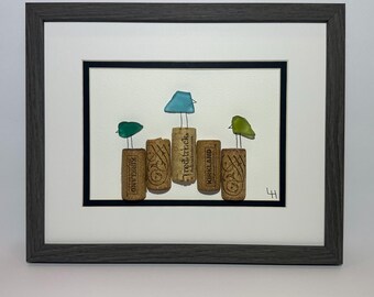 May include: Framed artwork featuring five wine corks with colorful glass bird toppers. The corks are arranged in a row, with green, blue, and yellow birds perched on top. The artwork is framed in a gray wooden frame.