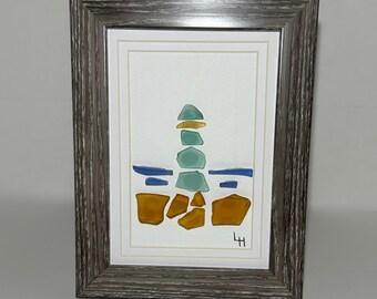 May include: Framed artwork featuring a lighthouse design made of sea glass. The lighthouse is composed of stacked teal, yellow, and blue glass pieces. The artwork is set against a white background and framed in a gray wooden frame.