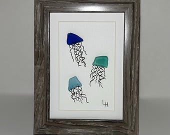 May include: Framed artwork featuring three jellyfish in shades of blue and green. The jellyfish are depicted with detailed, flowing tentacles against a white background. The artwork is encased in a gray wooden frame, adding a rustic touch.