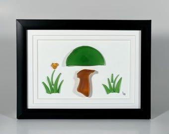 May include: Framed artwork featuring a green glass mushroom with a brown stem and a yellow flower, surrounded by green glass blades of grass. The artwork is set against a white background and framed in black.