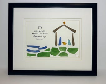 Könnte beinhalten: Gerahmtes Kunstwerk mit einer Krippenszene aus farbigem Meeresglas. Das Bild zeigt einen Stall, Figuren und eine Steinmauer. Der Text lautet "we love because he first loved us" aus 1. Johannes 4:19.