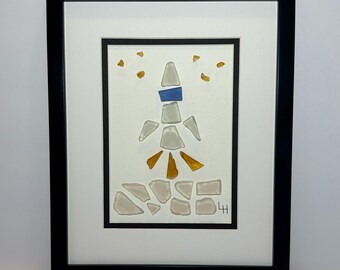 May include: Framed artwork featuring a mosaic design of a figure made from sea glass. The figure is composed of white, blue, and gold pieces, with a black frame and white matting. The artwork is a unique, handcrafted piece.
