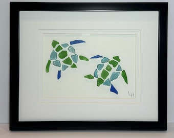 May include: Framed artwork featuring two sea turtles. The turtles are crafted from sea glass in shades of green, blue, and aqua, set against a white background. The artwork is encased in a black frame, creating a striking contrast.