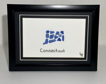 May include: Framed artwork featuring a white background with a blue mosaic of the state of Connecticut. The word "Connecticut" is written below the mosaic in black. The frame is black.