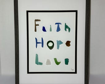 May include: Framed artwork with the words "Faith Hope Love" spelled out in colorful, irregularly shaped glass pieces. The artwork is framed in a gray wooden frame with a white mat.