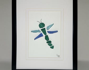 May include: Framed artwork featuring a dragonfly design. The dragonfly is crafted from various shades of blue and green glass pieces, set against a white background. The artwork is encased in a black frame, adding a modern touch to the piece.