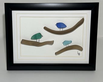 May include: Framed artwork featuring three colorful birds perched on brown branches. The birds are in shades of blue and green, set against a cream-colored background. The artwork is framed in a black frame.