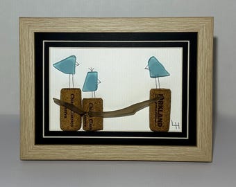 May include: A framed artwork featuring three light blue glass birds perched on wine corks. A brown glass branch connects the corks. The artwork is set against a white background with a black border, all within a light wood frame.