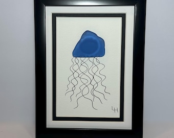 May include: Framed art print featuring a blue jellyfish design. The jellyfish has a rounded top and wavy black tentacles. The artwork is set against a white background, framed in a black frame. The artist's initials are in the lower right corner.