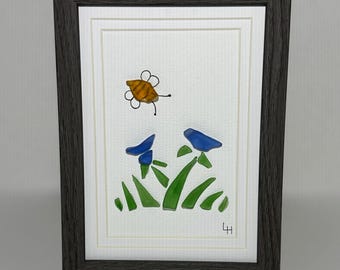 May include: Framed artwork featuring a bee and blue flowers. The bee is yellow and brown, and the flowers are blue. Green grass is at the bottom. The frame is dark gray.