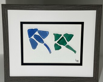 May include: Framed artwork featuring abstract designs made of blue and green sea glass. The sea glass pieces are arranged on a white background, framed with a black mat, and a gray wooden frame. The artwork is signed in the lower right corner.