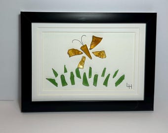 May include: Framed artwork featuring a flower and grass design made from sea glass. The flower has amber-colored petals and a black wire stem and antennae. Green sea glass pieces form the grass. The artwork is set against a white background and framed in black.