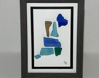 May include: Abstract art piece in a dark gray frame. The artwork features a collection of colorful glass pieces in shades of blue, brown, and green, arranged on a white background. The artist's initials are in the lower right corner.