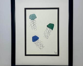 May include: Framed artwork featuring three jellyfish in shades of blue and green. Each jellyfish has a bell-shaped top and delicate, wavy tentacles. The artwork is framed in a black frame with a white mat.