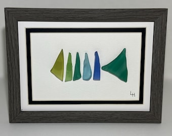 May include: Framed artwork featuring a fish skeleton design. The artwork uses various shades of green, blue, and yellow glass pieces arranged on a white background. The frame is a dark gray color, with a white matting and a black border.