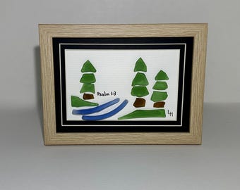 May include: Framed artwork featuring a minimalist landscape with green trees, brown accents, and blue water. The artwork includes the text "Psalm 1:3" and is set within a black mat and a light wood frame.