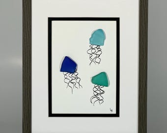 May include: Framed artwork featuring three jellyfish made from sea glass in shades of blue and green. The jellyfish are arranged in a cluster on a white background with black ink outlines.