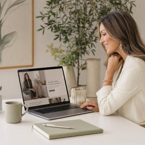 Peut inclure: Un ordinateur portable argenté affichant un site web intitulé Personal Coaching For Professional Women, posé sur un bureau blanc avec une tasse vert sauge, un carnet vert sauge assorti et un stylo argenté. L'écran montre une photo en costume vert.