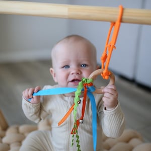 May include: A baby in a beige onesie holds a wooden ring with colorful ribbons and a blue plastic piece. The ring is suspended from a wooden play gym with orange straps. The baby has blue eyes and is looking at the camera.